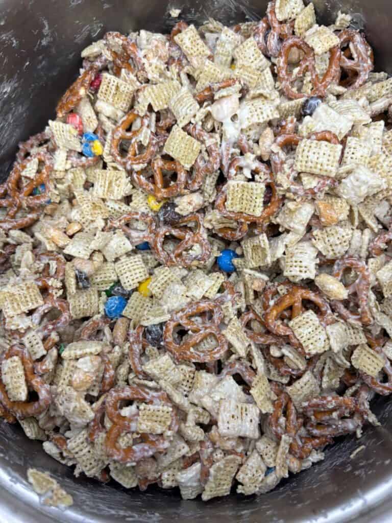 A close-up of a snack mix with pretzels, Chex cereal, MMs, and nuts coated in a white sugary glaze, all mixed together in a large metal bowl.