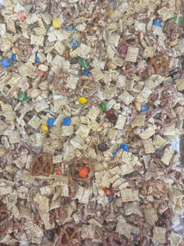 A close-up of a snack mix with pretzels, Chex cereal, MM candies, peanuts, and a white coating.