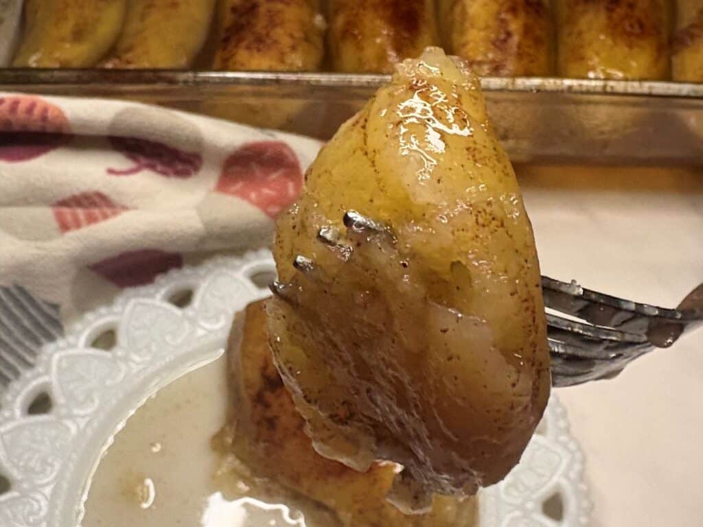 A fork holding a glazed, cooked banana slice above a small plate with creamy sauce.
