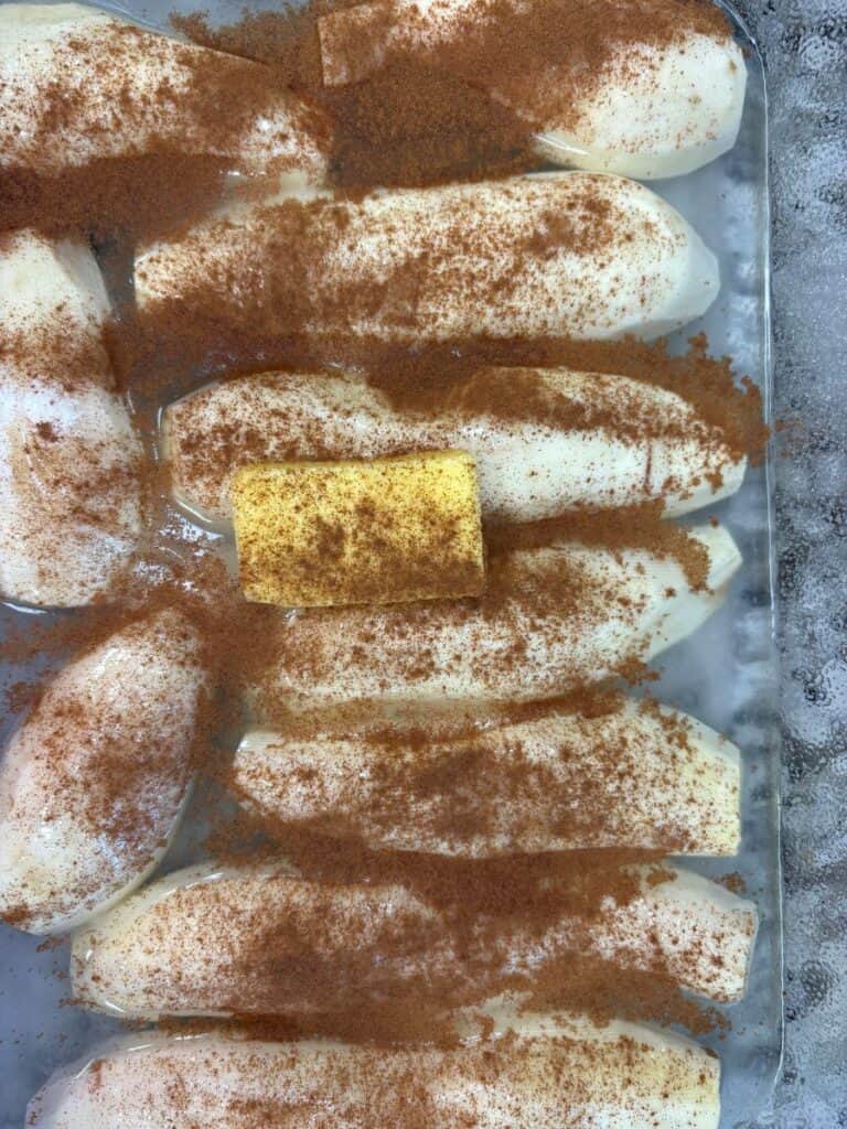 Peeled sweet potato halves arranged in a glass baking dish, dusted with cinnamon and topped with a pat of butter.