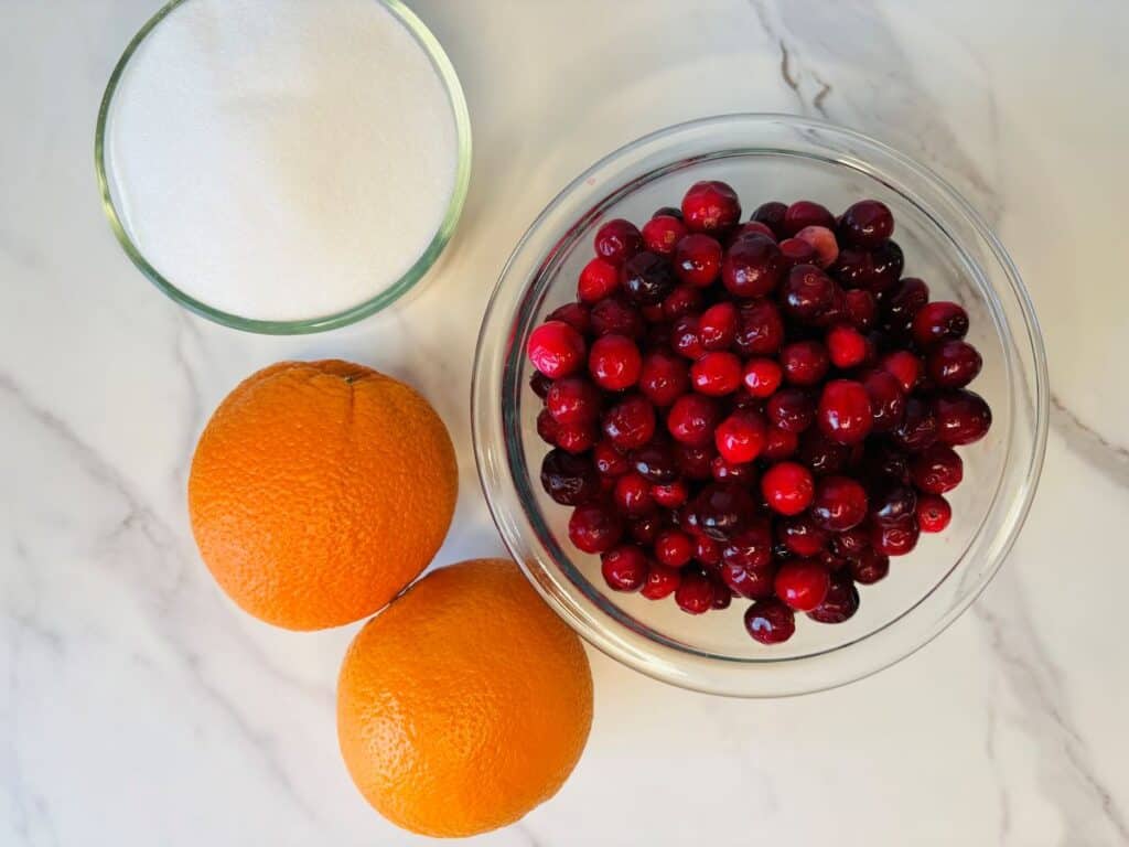 A bowl of fresh cranberries, two whole oranges, and a small bowl of granulated sugar.