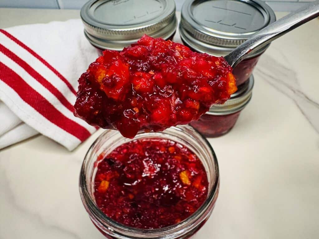 A spoonful of bright red cranberry orange relish held over an open glass jar, with two additional sealed jars in the background.