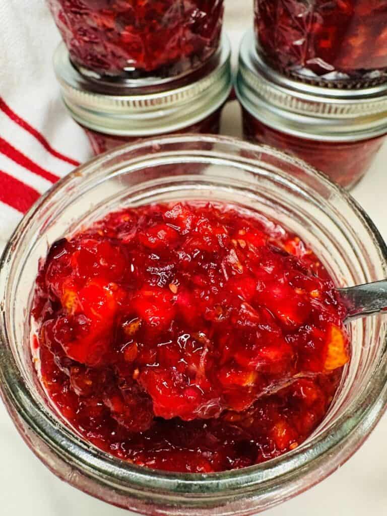 A spoonful of cranberry orange relish held over an open glass jar, with additional jars in the background.