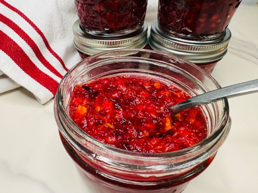 A metal spoon resting in an open glass jar of chunky red cranberry sauce, with other sealed jars and a striped towel nearby.