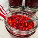 A metal spoon resting in an open glass jar of chunky red cranberry sauce, with other sealed jars and a striped towel nearby.