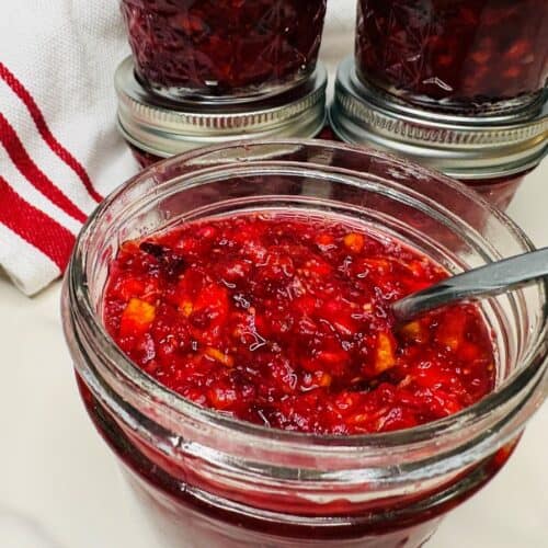 A metal spoon resting in an open glass jar of chunky red cranberry sauce, with other sealed jars and a striped towel nearby.