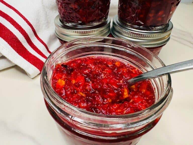 A metal spoon resting in an open glass jar of chunky red cranberry sauce, with other sealed jars and a striped towel nearby.