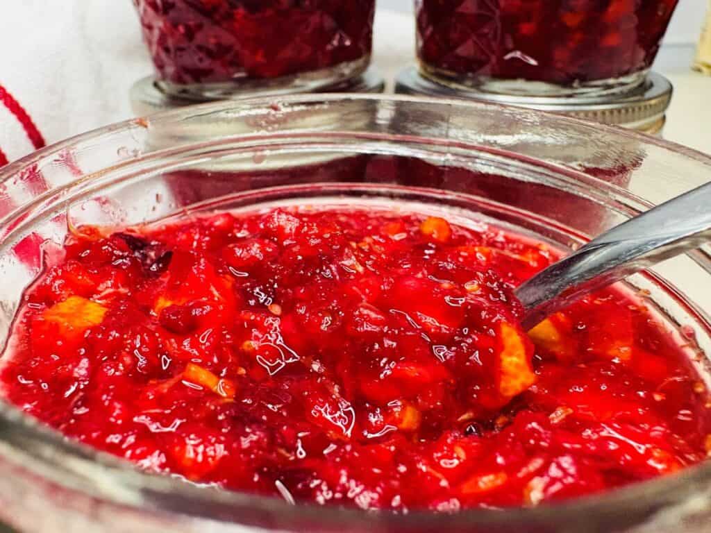A glass jar filled with red fruit preserves and a metal spoon, with two additional jars of preserves in the background.