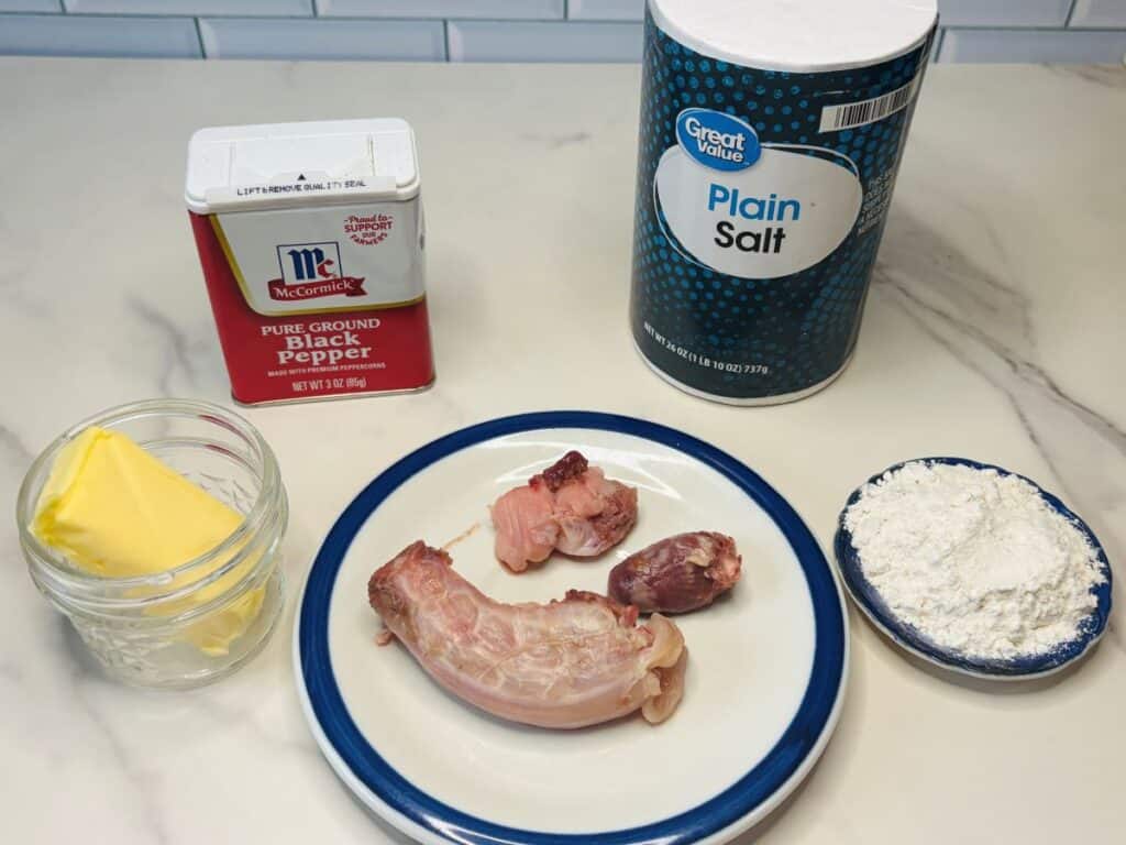 Ingredients for a recipe: a jar of butter, black pepper, salt, flour in a bowl, and a plate with a raw chicken neck, liver, and gizzard on a white countertop.