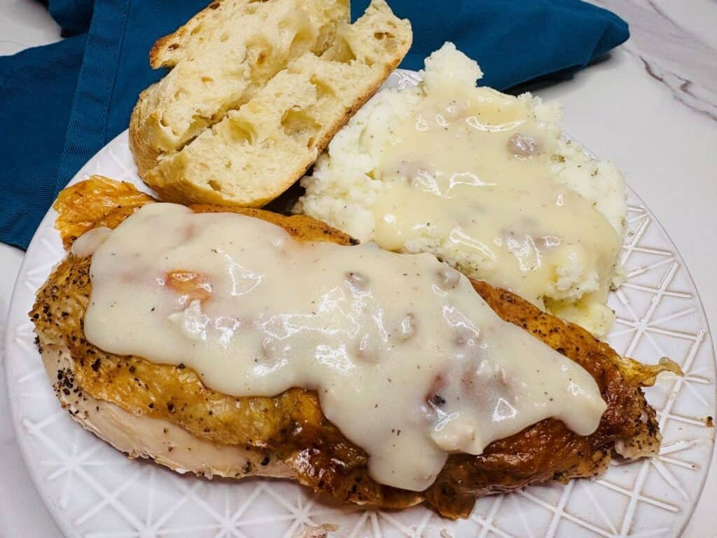 A plate with roasted chicken topped with white gravy, mashed potatoes with gravy, and two slices of rustic bread. A blue napkin is beside the plate on a marble surface.