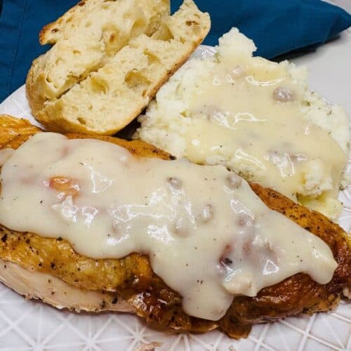 A plate with roasted chicken topped with white gravy, mashed potatoes with gravy, and two slices of rustic bread. A blue napkin is beside the plate on a marble surface.