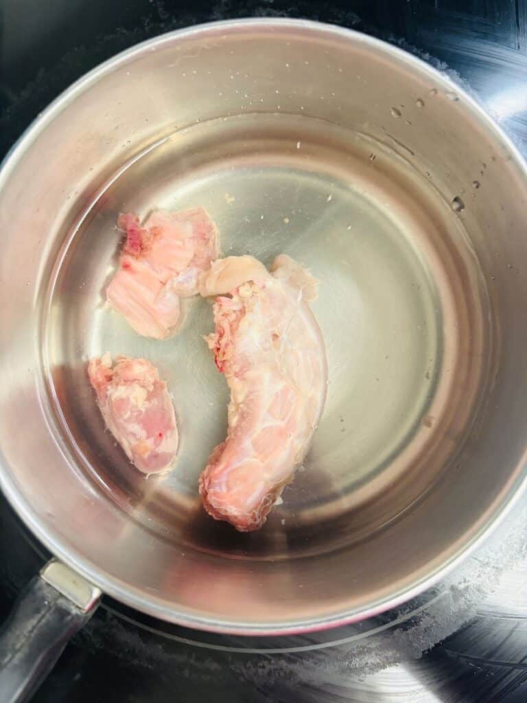 Raw pieces of chicken placed in a stainless steel pot filled with water, sitting on a stovetop, ready to be boiled or cooked.