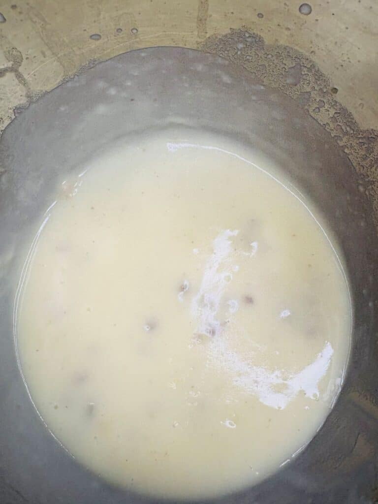 A close-up of a thick, creamy, off-white liquid with small lumps in a metal pot, possibly a soup or sauce, with residue visible on the sides.