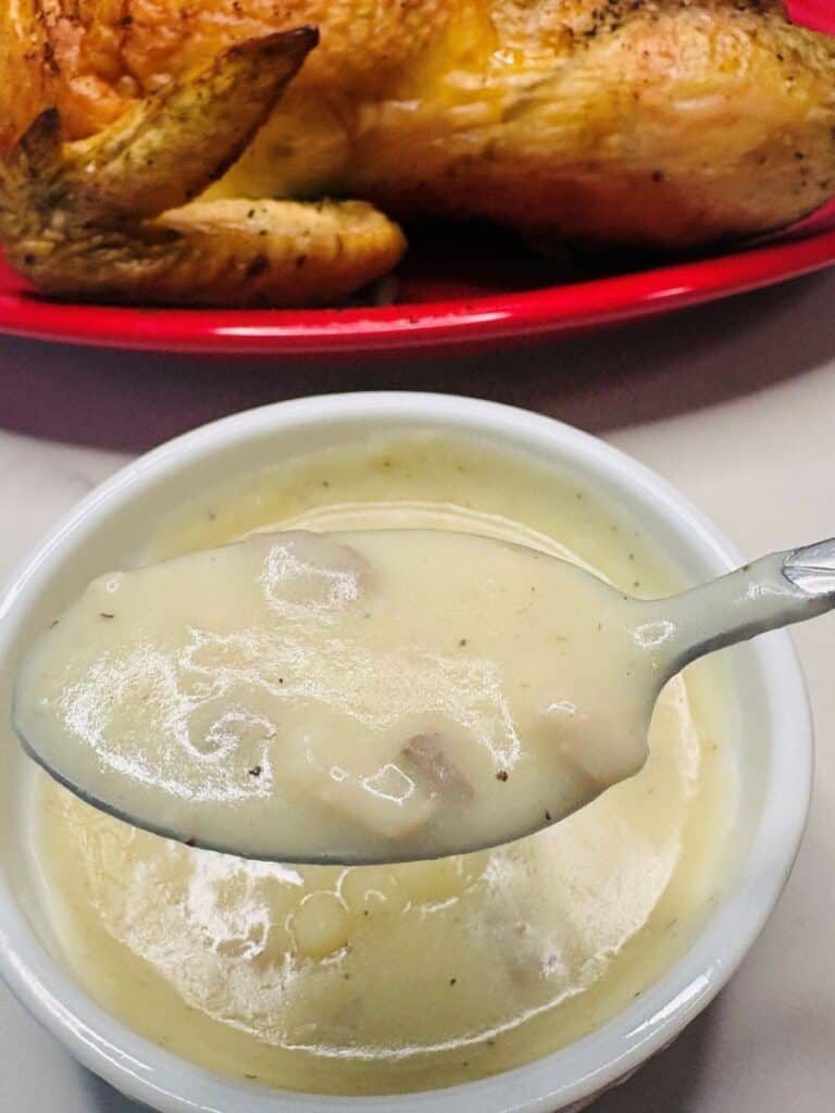 A close-up of a spoonful of creamy gravy with visible bits in it, held above a small white bowl. In the background, part of a roasted chicken on a red plate is visible.