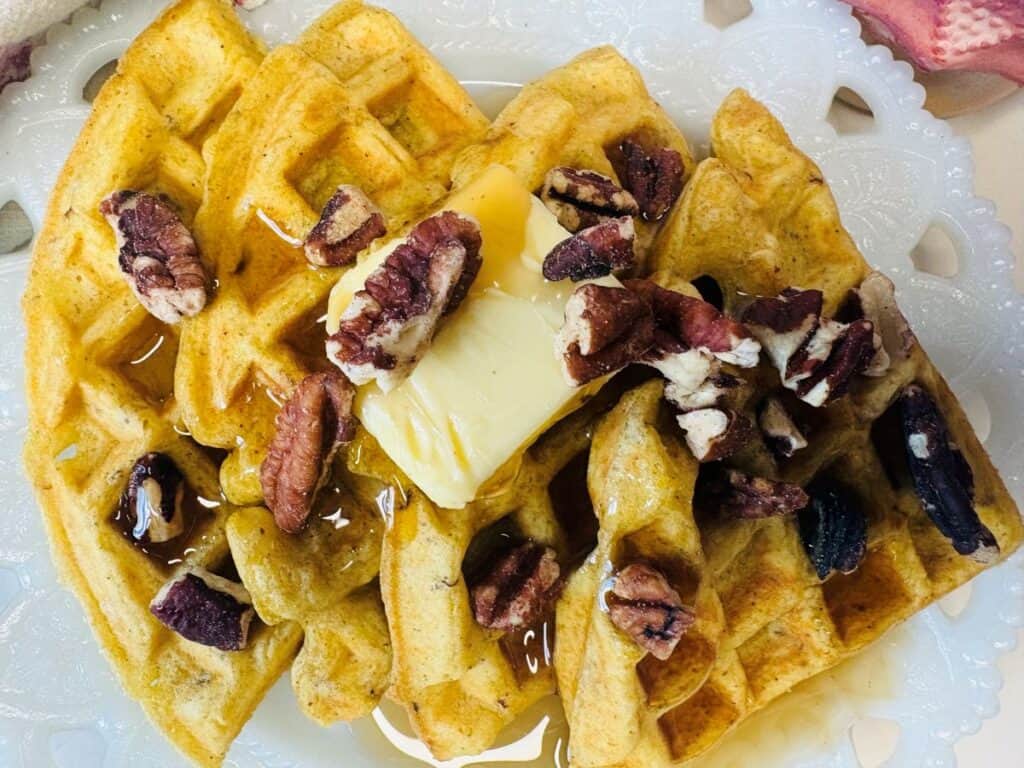 Golden waffles topped with a pat of butter, pecans, and syrup on a white decorative plate.