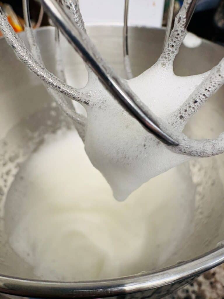 Wire whisk of a stand mixer lifting foamy, whipped egg whites from a stainless steel mixing bowl.