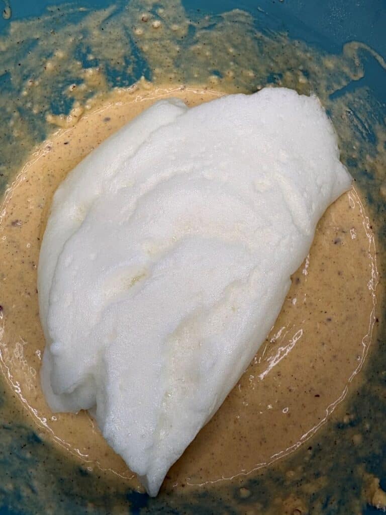 A mound of fluffy, white whipped egg whites resting on top of a tan, speckled cake batter in a blue bowl.