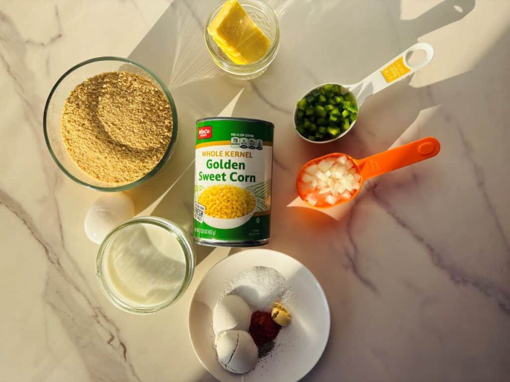 Ingredients for a recipe, including a can of golden sweet corn, breadcrumbs, milk, an egg, butter, chopped green peppers, onions, and small piles of spices on a marble surface.