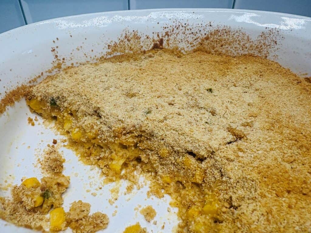 A baked corn casserole in a white ceramic dish with a golden-brown breadcrumb topping and a large serving scooped out.